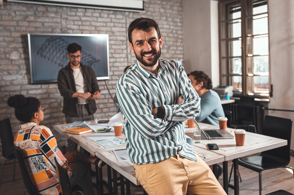 Portrait of confident young male leader standing and looking at camera while her colleagues working on architectural designs in background